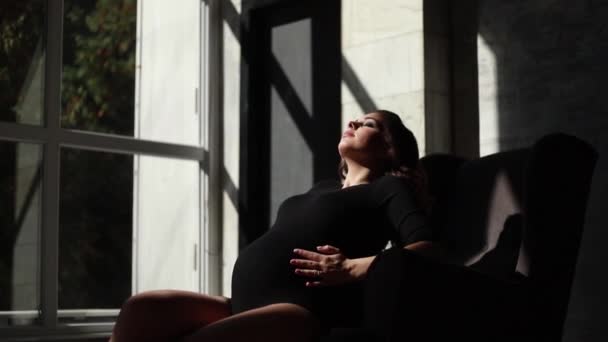 Красивая беременная женщина сидит на стуле. Солнечные лучи падают на женщину. Она одета в чёрный костюм. — стоковое видео