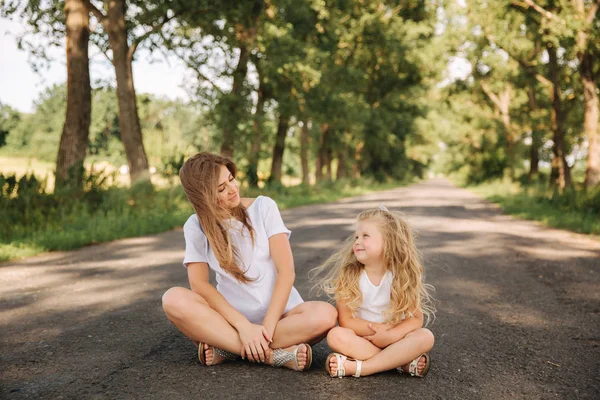 Attractive Mom and blonde hair daughter sits on road near big alley. They smile and look to natune. Front view — Stock Photo, Image