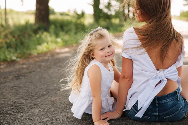 Attractive Mom and blonde hair daughter sits on road near big alley. They smile and look to natune. Front view — Stock Photo, Image