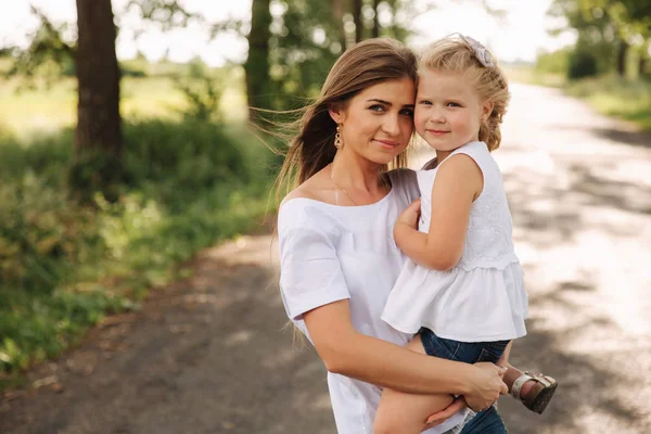 Attractive Mom and blonde hair daughter sits on road near big alley. They smile and look to natune. Front view