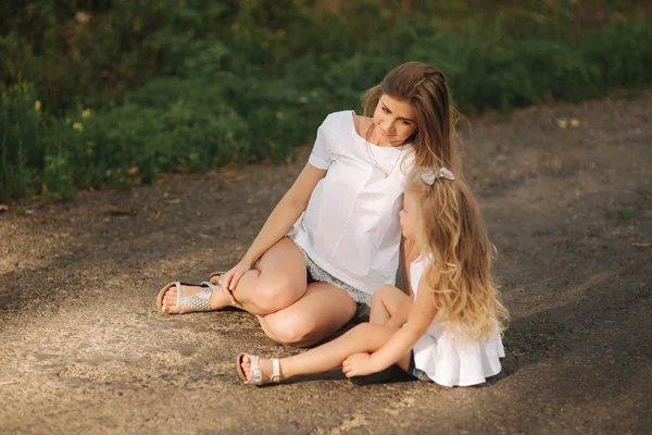 Attraktive Mutter und blonde Haare Tochter sitzt auf der Straße in der Nähe der großen Gasse. sie lächeln und schauen zur Natur — Stockfoto