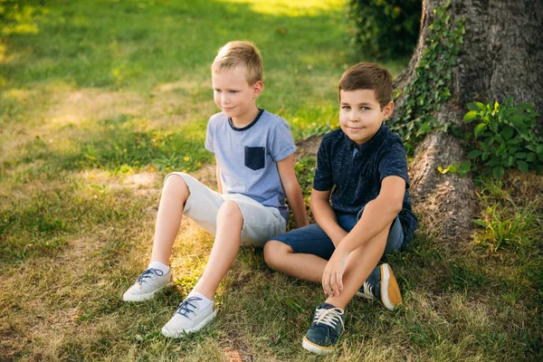 Dos chicos se sientan en la hierba cerca del árbol. Niños de diez años pasan tiempo en el parque —  Fotos de Stock