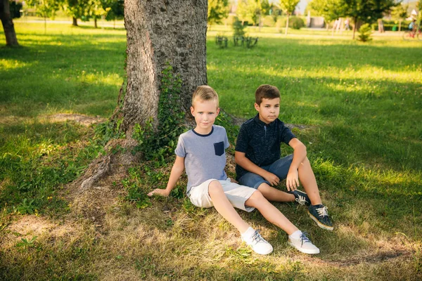 Dos chicos se sientan en la hierba cerca del árbol. Niños de diez años pasan tiempo en el parque —  Fotos de Stock