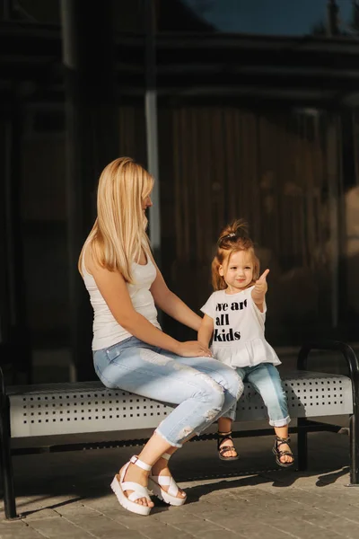Mother and daughter outdoors in city. playing and having fun. Fashion happy mother and child daughter having fun together in the city. They are sitting on the bench near the mall — Stock Photo, Image