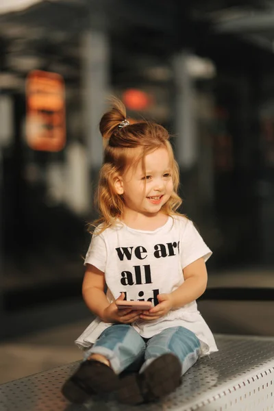 Little girl is sitting on benches and using phone near the mall — Stock Photo, Image