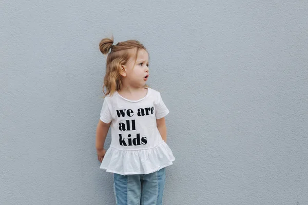 아름 다운 작은 소녀 회색 벽의 앞에 흰색 셔츠와 청바지 스탠드 입고. 행복 한 아이 들 — 스톡 사진