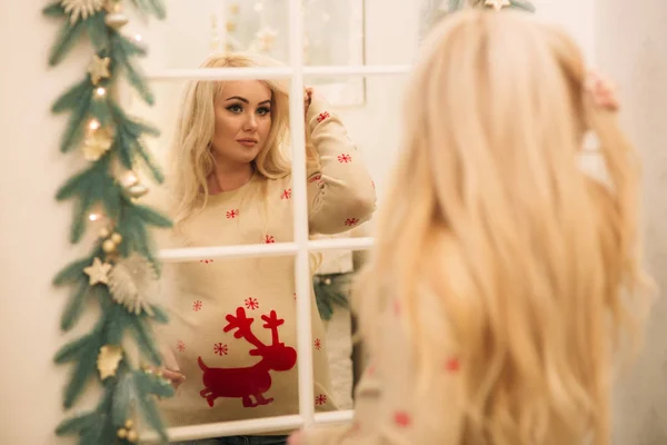 A pretty pregnant blonde hair woman in a Christmas sweater stands near the mirror. Winter spirit. Happy New Year