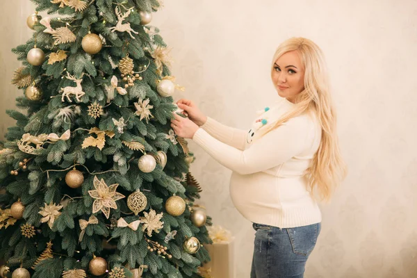 En ganska gravid blondin i jul tröja står nära julgran — Stockfoto