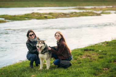 İki kız kardeş ile nehir önünde husky köpek yürümek. Brunettle ve sarışın kız bir köpek okşamak