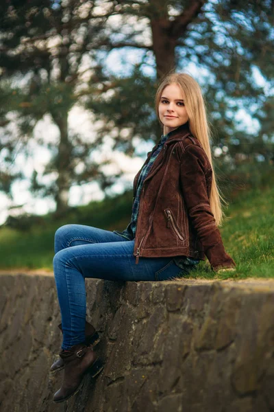 Aantrekkelijke jonge meisje door de dijk zit. Blond haar en bruine jas. Lente — Stockfoto