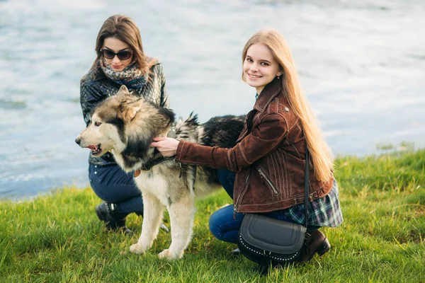 Two sisters walk with husky dog in front of river. Brunettle and blonde girl caress a dog — Stock Photo, Image