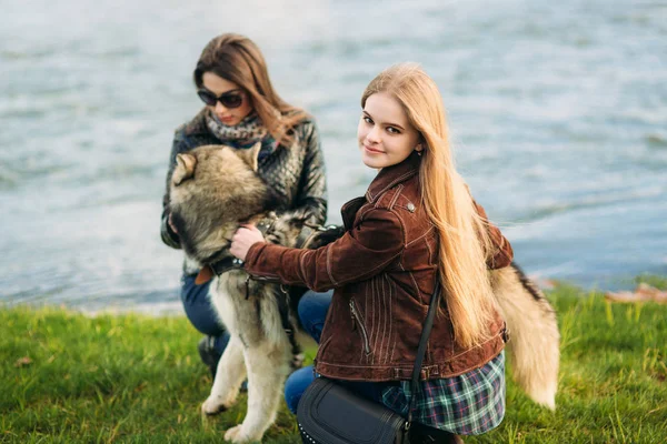 Zwei Schwestern spazieren mit Husky-Hund vor dem Fluss. Brünette und blonde Mädchen streicheln einen Hund — Stockfoto