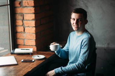 Yakışıklı çocuk kafede kahve içme. Pencerenin laptop ve defter ile oturuyor