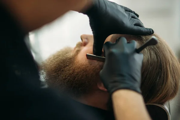 Bearded man with long beard getting stylish hair shaving , haircut , with razor by barber in barbershop