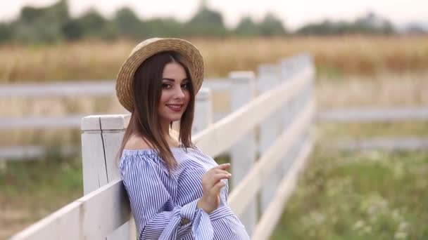 La mujer embarazada en la naturaleza se prueban el sombrero de punto. Antecedentes de campo y fachada blanca cerca de la granja — Vídeos de Stock