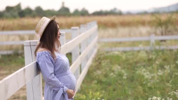 La mujer embarazada en la naturaleza se prueban el sombrero de punto. Antecedentes de campo y fachada blanca cerca de la granja — Vídeos de Stock