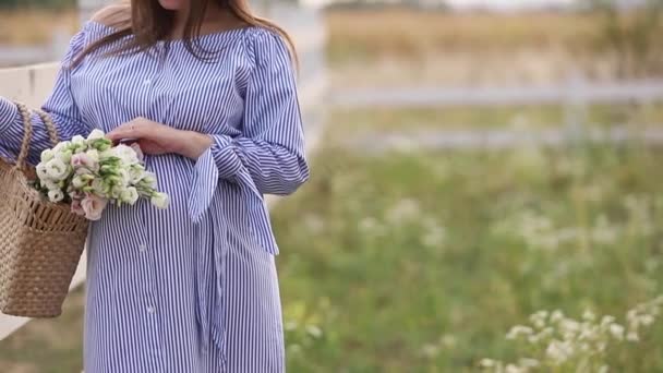 Beautiful pregnant woman in blue dress walking hear the farm. Knitted bag with bouquet of flowers. Background of tree and mountains. Slow motion