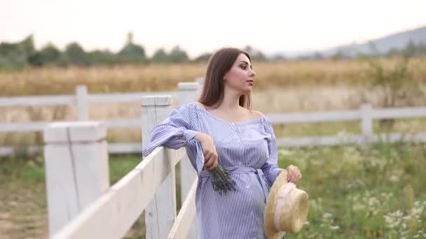 Беременная женщина в природе примеряет вязаную шляпу. Фон поля и белый забор возле фермы — стоковое видео