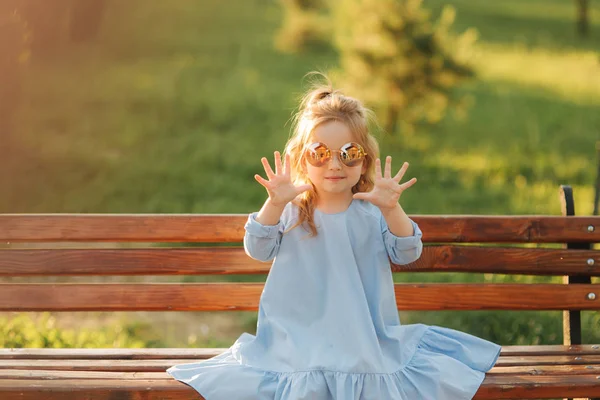 Маленькая девочка модель в синем платье и солнцезащитных очках сидит на скамейке в парке — стоковое фото