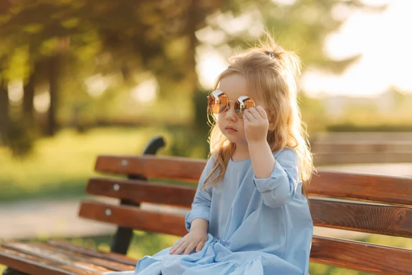 Маленькая девочка модель в синем платье и солнцезащитных очках сидит на скамейке в парке — стоковое фото