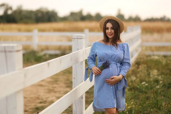 Hermosa mujer embarazada en vestido azul caminando cerca de la granja. Bolso de punto y ramo de lavanda. Contexto de la explotación —  Fotos de Stock