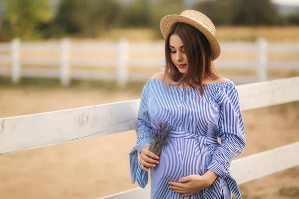 Hermosa mujer embarazada en vestido azul caminando cerca de la granja. Bolso de punto y ramo de lavanda. Contexto de la explotación —  Fotos de Stock