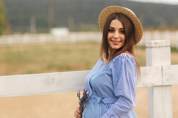 Красива вагітна жінка в блакитній сукні, що йде поблизу ферми. В'язаний мішок і букет лаванди. Фундамент ферми — стокове фото