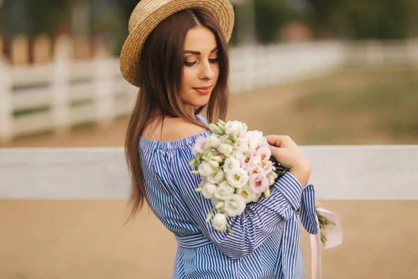 Eine Frau steht vor einem Bauernhof und hält einen Blumenstrauß in den Händen. schöner Strauß. Frau in blauem Kleid und Strickmütze — Stockfoto