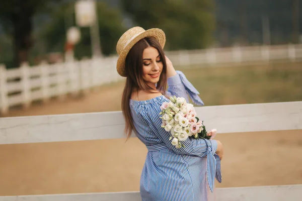Georgeus femme debout devant la ferme et tenir un bouquet de fleurs dans les mains. Magnifique bouquet. Femme en robe bleue et chapeau tricoté — Photo