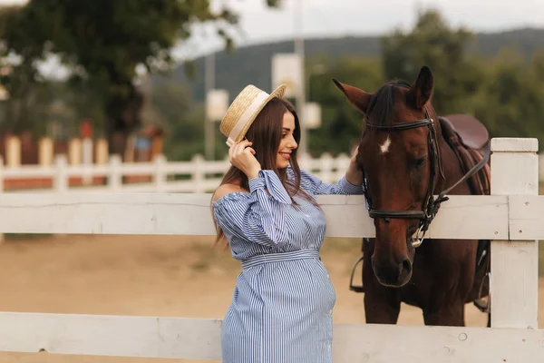 Mulher grávida bonita ficar perto do cavalo. Senhora de chapéu de malha e vestido azul. Bonito animal de quinta. Brown Hors — Fotografia de Stock