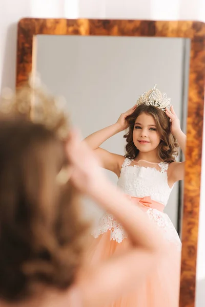 Kleines schönes Mädchen mit braunen Haaren in einem pfirsichfarbenen Kleid. Mädchen probieren Diadem an. glückliche kleine Prinzessin schaut in den Spiegel — Stockfoto