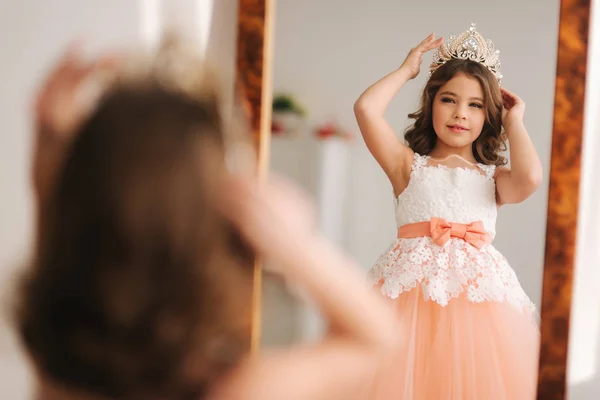 Little beautiful girl with brown hair in a Peach-colored dress. Girl try on diadem. Happy little princess look in to the mirror