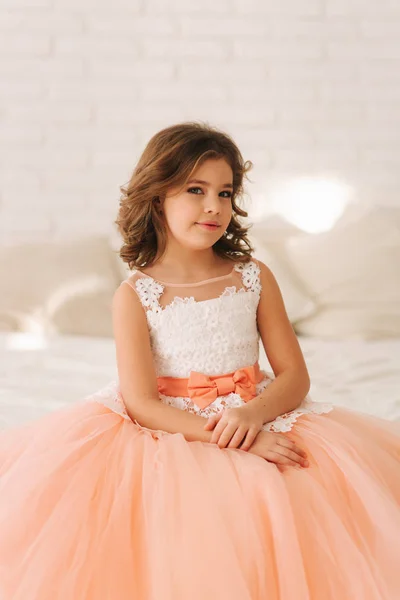 Маленькая красивая девочка с каштановыми волосами в персиковом платье. Девушка сидит на диване — стоковое фото