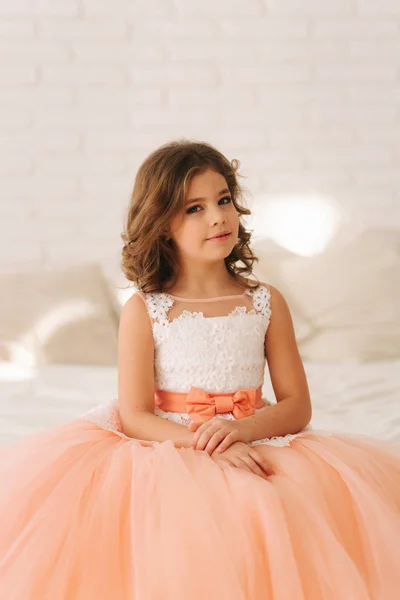 Little beautiful girl with brown hair in a Peach-colored dress. Girl sitting on the sofa