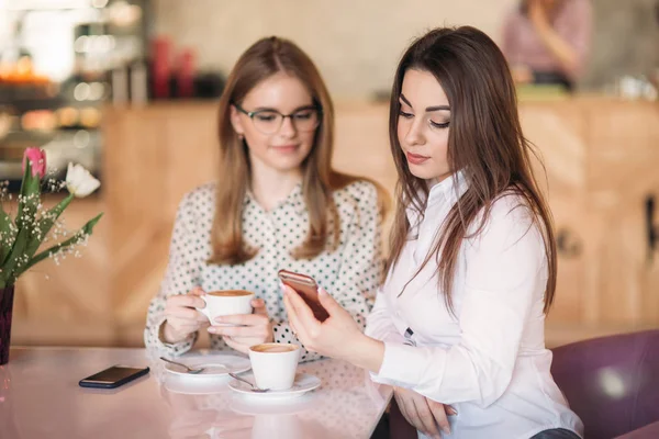 Two girls sitting in a cafe drinking cappuccino and using smartphone. Modern girl — Stock Photo, Image