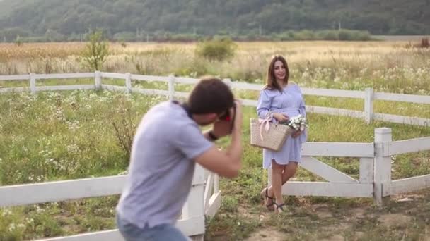 Fotograf zrobić zdjęcie dla piękna w ciąży kobieta ubrana w niebieską sukienkę i czapka. Położyła ręce na jej brzuch. Photosession w polu w pobliżu gospodarstwa. Czas letni — Wideo stockowe