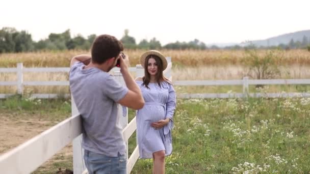 Fotoğrafçı mavi elbiseli giyinmiş ve şapka örme güzel hamile kadın için bir fotoğraf olun. Karnı üzerinde el koymak. Photosession farm yakınındaki alanda. Yaz saati — Stok video