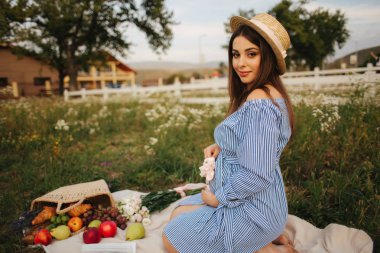 Sevimli hamile kadın te fieln ekose üzerinde oturuyor. Kadın mavi elbise giymiş ve şapka örme. Bir meyve sepeti götürdü. Çiftlik arka plan