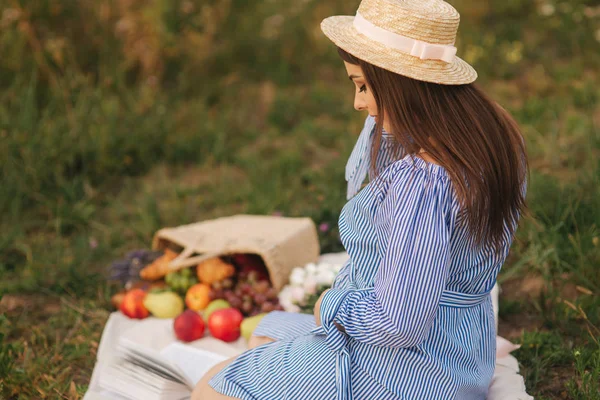 Mujer embarazada joven sentada en el campo y relajarse. Se llevó una cesta de frutas. Verano — Foto de Stock