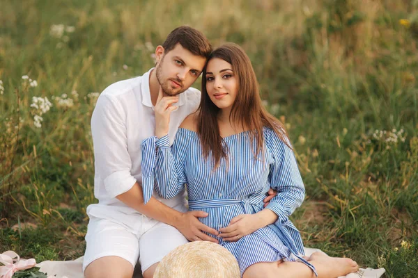 Die zukünftige Mutter und der zukünftige Vater sitzen auf dem Feld. Schwangere mit ihrem Mann legte die Hände auf den Bauch — Stockfoto