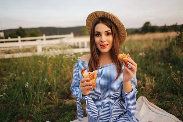 Mujer embarazada en el campo comiendo croissant crujiente respaldado fresco. Comida saludable. La joven come muy apetitoso. Contexto de la explotación — Foto de Stock