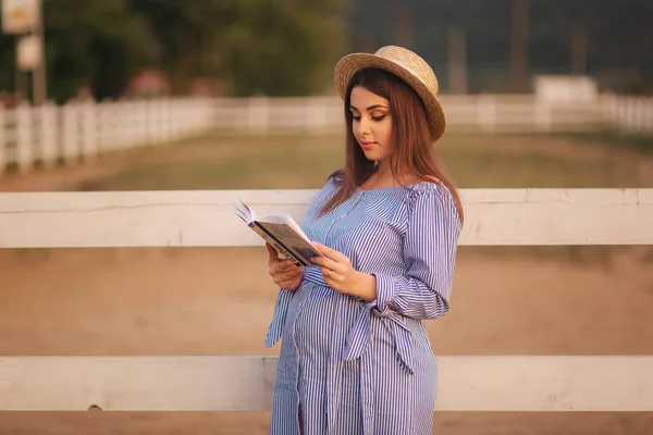 Beautiful pregnant woman read the book on the farm. She stand by the fance and look in to the book. Relax