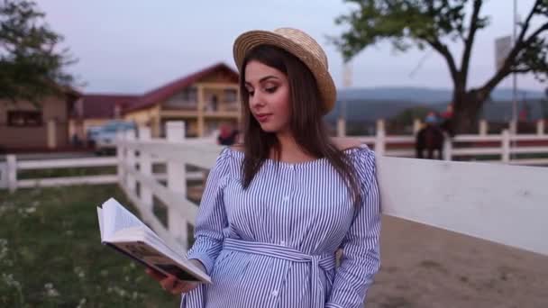 Mulher grávida bonita ler o livro na fazenda. Ela fica ao lado da cerca e olha para o livro. Relaxa. — Vídeo de Stock