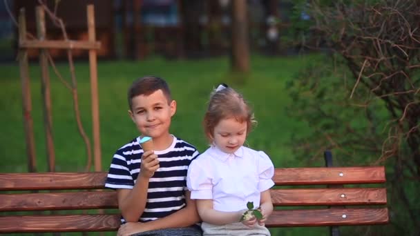 Mały chłopiec i dziewczynka siedzi na ławce i zjada lody — Wideo stockowe
