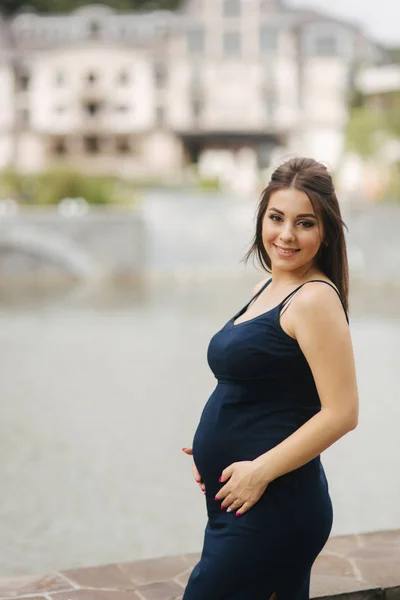 Красива молода весела вагітна жінка, що стоїть на фоні фонтану. шість місяців вагітності. Вона наклала руки на живіт — стокове фото