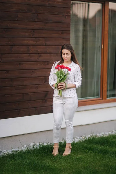 Красива дівчина з букетом червоних квітів стоїть біля будинку. Дерев'яний фон — стокове фото