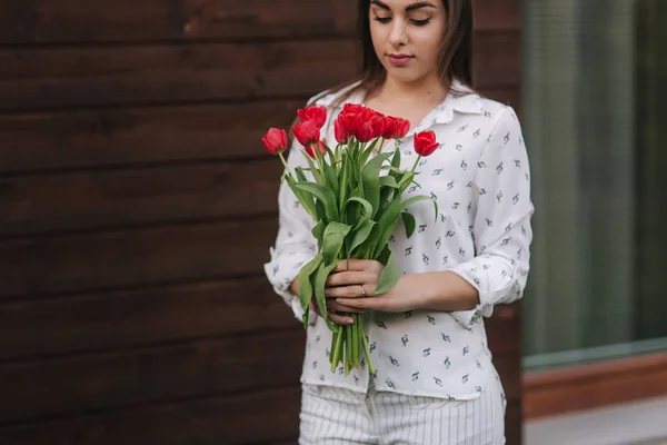 Красива дівчина з букетом червоних квітів стоїть біля будинку. Дерев'яний фон — стокове фото