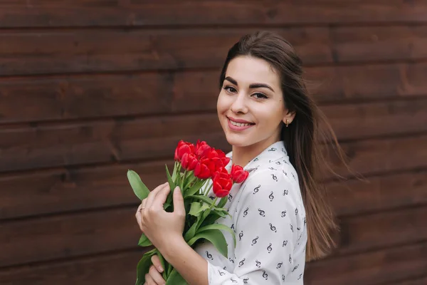 Ein junges Mädchen mit einem Strauß roter Tulpen steht draußen vor einem hölzernen Hintergrund. glückliches Weibchen bekam einen Blumenstrauß — Stockfoto