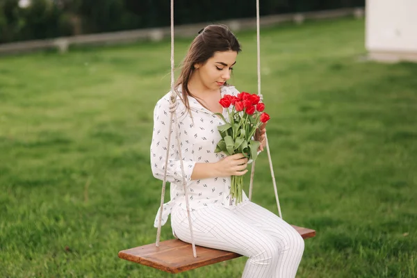 La señora de blanco se sienta en el columpio en el patio trasero de su casa. Ella sostiene una re flores —  Fotos de Stock