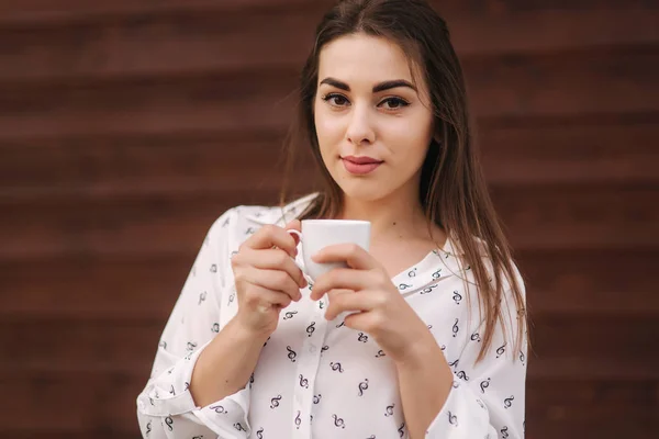 Gyönyörű lány tart egy csésze kávét. Fehér inget. Fából készült háttér — Stock Fotó